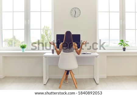 Back view of woman taking break from office work and meditating sitting in modern workspace at table with desktop computer and cup of coffee to go. Concepts of corporate wellbeing and stress relief