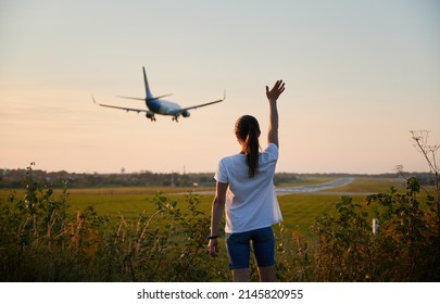 Back View Of Woman Waving Hand To Landing Commercial Airplane At The Airport In The Evening. Lifestyle And Travel Concept.