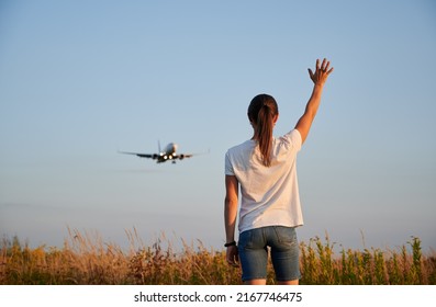 Back View Of Woman Waving Hand To Flying Commercial Airplane In The Sky At Sunset. Lifestyle And Travel Concept.