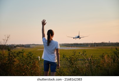 Back View Of Woman Waving Hand To Landing Commercial Airplane At The Airport In The Evening. Lifestyle And Travel Concept.