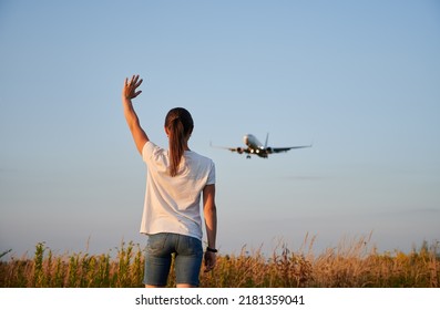 Back View Of Woman Waving Hand To Flying Commercial Airplane In The Sky In The Evening. Lifestyle And Travel Concept.