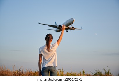 Back View Of Woman Waving Hand To Flying Commercial Airplane In The Sky At Sunset. Lifestyle And Travel Concept.