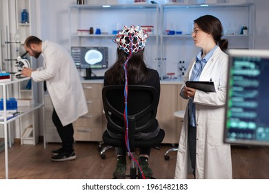 Back View Of Woman Wearing Headset For Brain Scan In Neurology Laboratory, Neuro Science. Nervous System Analysed By Team Of Scientists. Medical Scientist Looking Through Microscope In Background