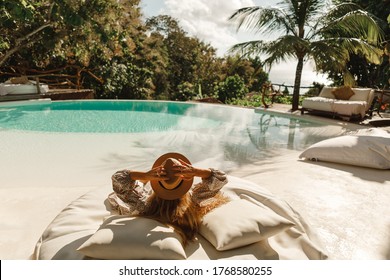 Back view woman wearing straw hat lie on sunbed  and enjoy sun tan near swimming pool with view on ocean and palm trees.  Relaxing summer day, Luxurious tropical vacation concept - Powered by Shutterstock
