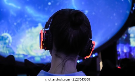 Back view: woman wearing wireless black headphones and looking around in dark room of interactive exhibition or museum with colorful illumination. Futuristic, entertainment, immersive concept - Shutterstock ID 2127643868