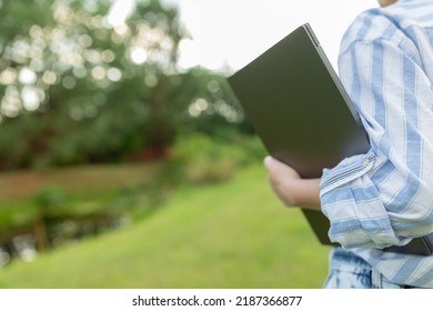 Back View Of A Woman Who Is Standing With A Closed Laptop. Rear View People Collection. Backside View Of Person. Young Engineer With A Closed Laptop In Hand.