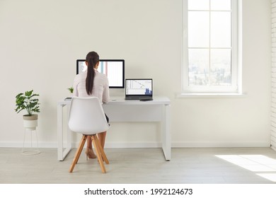 Back View Of A Woman Working In The Interior Of A Clean Spacious Office. Young Female Worker Working With Spreadsheets Sitting At A Desk With A Modern Desktop Computer And A Laptop