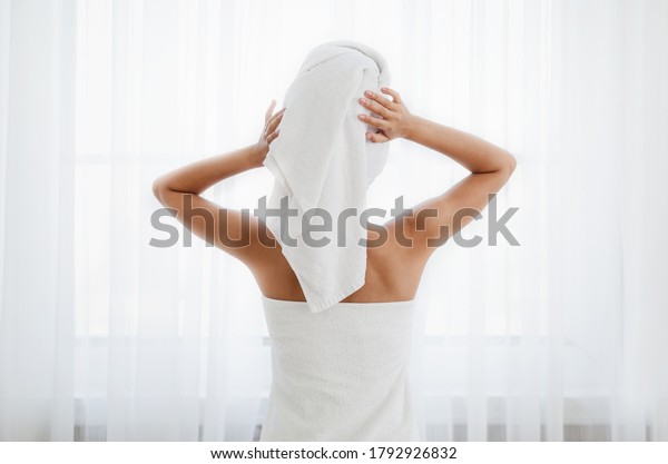 Back view of woman wrapped in\
towel standing next to window, drying her hair after morning\
shower