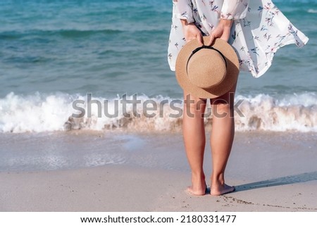 Back view of woman's legs standing on sandy beach near turquoise colored ocean in summertime. Female tourist with straw hat in hands enjoying traveling to exotic nature on a beautiful sunny day. 