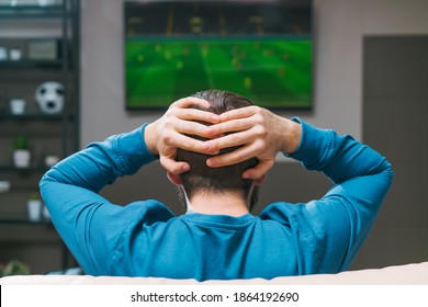 Back View Of Yong Bearded Man Watching Tv And Holding His Head With Hands Stressed About Football Play Result. He Lost His Bet.