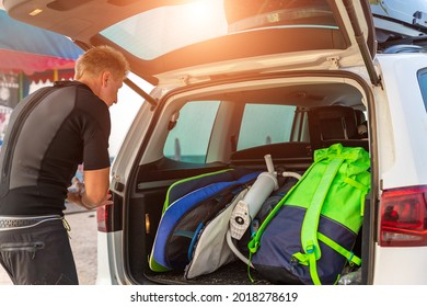 Back view young adult male surfer in wetsuit put out surf board kite equipment on sand beach from van vehicle with rooftop box ocean sea beach watersport spot camp.Adventure travel sport acitivity