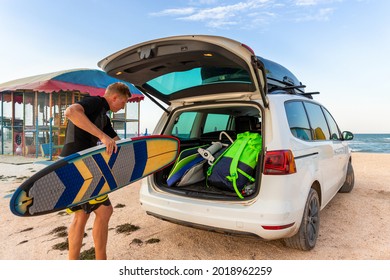 Back view young adult male surfer in wetsuit put out surf board kite equipment on sand beach from van vehicle with rooftop box ocean sea beach watersport spot camp.Adventure travel sport acitivity