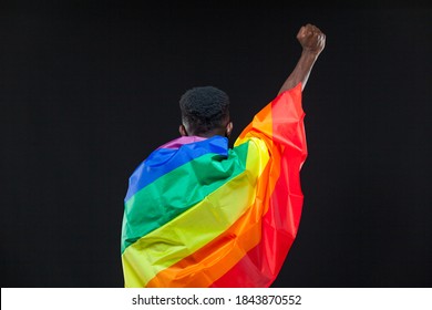 Back view of young african american man wrapped in a rainbow flag standing with raised fist isolated on black background. Concept of The LGBT community, minority rights, protection of human rights