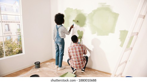 Back View Of Young African American Married Couple Man And Woman Working In Room Renovating Own House. Wife And Husband Painting Walls In Green Color Together Using Roller Brushes. Home Repair Concept