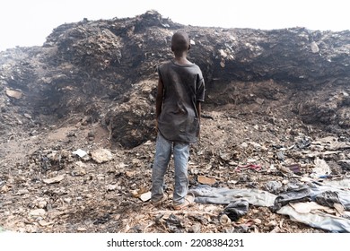 Back View Of A Young African Street Boy Facing A Huge Mountain Of Smoking Waste In An Urban Slum Area; Symbol Of Extreme Poverty In Least Developed Countries