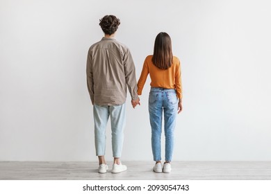 Back view of young Asian couple in casual wear standing, holding hands against white studio wall. Affectionate millennial girlfriend and boyfriend posing together, full length