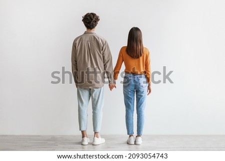 Back view of young Asian couple in casual wear standing, holding hands against white studio wall. Affectionate millennial girlfriend and boyfriend posing together, full length