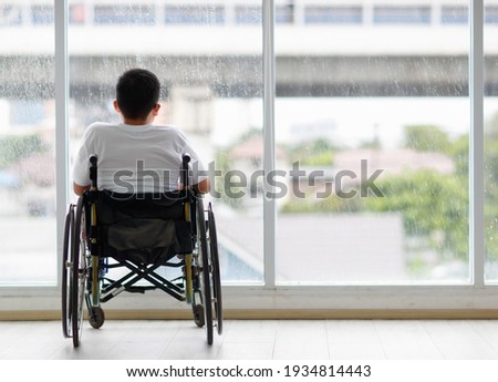 Back view of young Asian handicapped teenage boy without legs sitting on wheel chair and looking to outside of home with copy space on right.