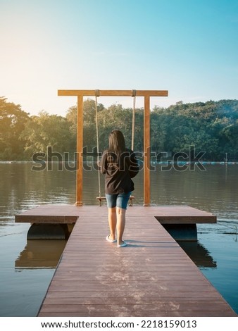 Back view of young Asian woman walking to play a wooden bench swings in summer holiday at bank of river. Lonely girl on waterfront alone and enjoying nature view at beautiful big river in evening.