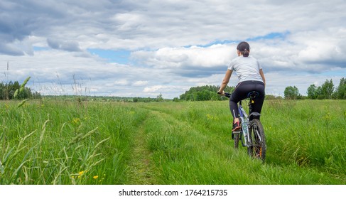 Back View Of Young Athletic Woman Riding Bicycle On Rural Road In Green Field Or Meadow In Summertime Against Dramatic Sky. Solo Outdoor Activities. Enjoy Time Alone In Nature. Banner, Copy Space