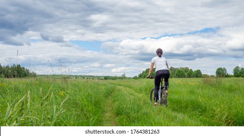 Back View Of Young Athletic Woman Riding Bicycle On Rural Road In Green Field Or Meadow In Summertime Against Dramatic Sky. Solo Outdoor Activities. Enjoy Time Alone In Nature. Banner, Copy Space