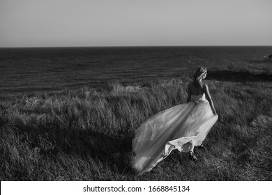 Back View Young Beautiful Caucasian Woman In Long Wedding Dress On Coast Of Sea. A Bride In A White Wedding Dress Standing On A Cliff Edge Looking Out To Sea. Black And White. Copy Space.