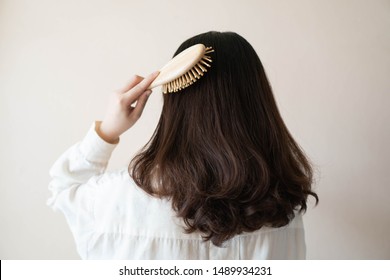 Back View Of Young Beautiful Woman In White Shirt With Long Black Curly Hair Combing Her Hair In The Morning. Hair Care Concept.