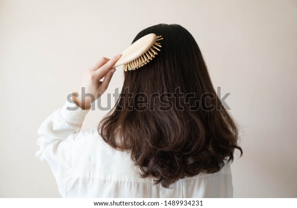 Back view of young beautiful woman in white\
shirt with long black curly hair combing her hair in the morning.\
Hair care concept.