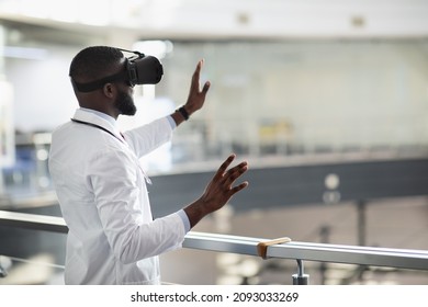 Back view of young black doctor in workwear and stethoscope using VR glasses, touching copy space, clinic interior, panorama. Modern technologies in healthcare, telemedicine concept