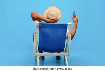Back view of young black guy sitting in lounge chair with bottle of beer, enjoying summer vacation, sipping alcoholic beverage, sunbathing on blue studio background, full length - Powered by Shutterstock