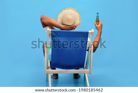 Back view of young black guy sitting in lounge chair with bottle of beer, enjoying summer vacation, sipping alcoholic beverage, sunbathing on blue studio background, full length