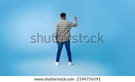 Back view of young black guy pushing button on virtual screen over blue studio background, mockup for design. Millennial African American man interacting with touchscreen, panorama
