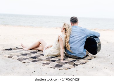 Back View Of Young Blonde Barefoot Woman Sitting On Checkered Blanket Near Boyfriend With Acoustic Guitar At Beach Near Sea