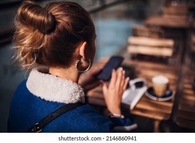 Back view of a young blonde woman drinking coffee in outdoors cafe. Texting a message on phone, photographing food on a mobile phone. Wearing stylish jeans coat. Lifestyle fashion portrait