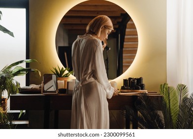 Back view of young blonde woman wearing robe talking on mobile phone while standing in front of mirror in bathroom - Powered by Shutterstock