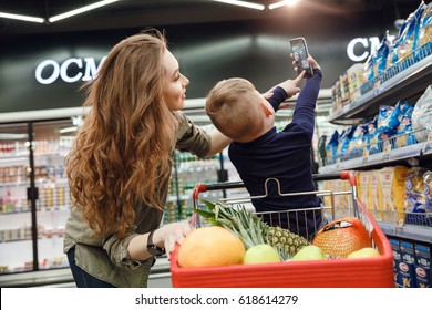 Back View Of A Young Boy Making Selfie With Mom On Smartphone In The Supermarket