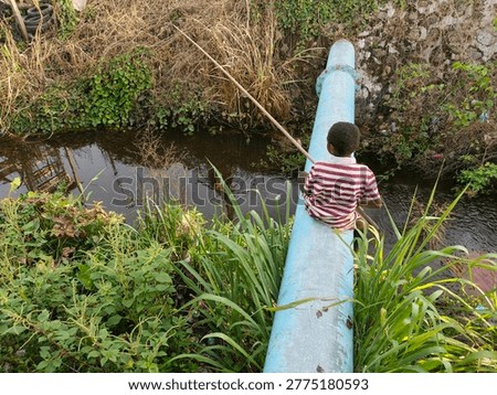 Back view of a young boy sitting on a metal water pipe fishing in a ditch with a traditional handmade rod in a rural village area