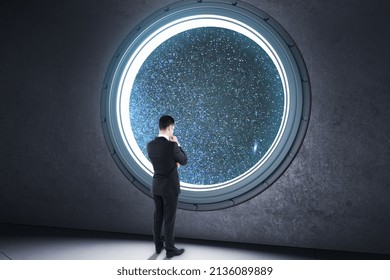 Back view of young business man looking out of round illuminator with starry sky cosmos view in concrete interior. Future and creativity concept