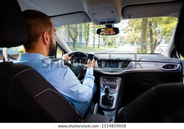 Back view of young businessman driving\
car and texting on mobile phone at the same time.\
