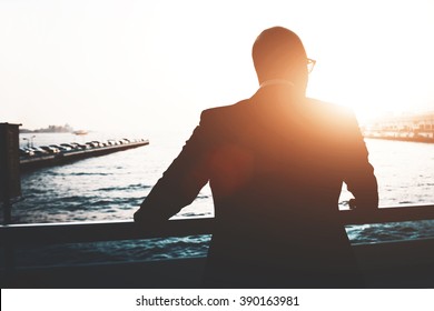 Back view of a young businessman is enjoying view and rest after meeting during his business trip. Silhouette of a male lawyer is resting after work day, while is standing outdoors against sunset - Powered by Shutterstock