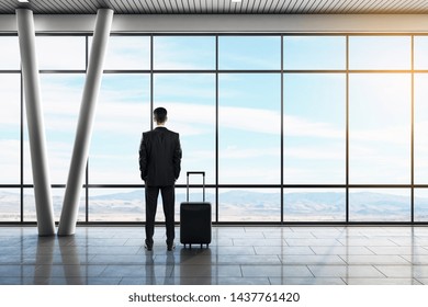 Back view of young businessman looking out of window in contemporary airport lounge interior with daylight, suitcase and airplane. Travel and vacation concept. - Powered by Shutterstock