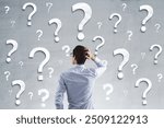 Back view of young businessman on concrete wall background with question marks. Solution and confusion concept