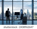 Back view of young businessman standing in modern office interior with empty computer monitor on desk and panoramic windows with beautiful city view and daylight. Mock up. Workplace concept