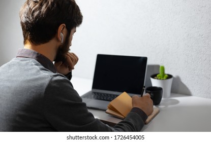 Back View Of Young Businessman Working Home At Quarantine, Making Notes In Notebook, Using Wireless Earphones, Laptop, Coffee Mug And Cactus On Work Desk. Background Of Textured Wall Of Grey Color.