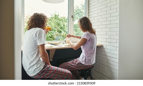 Back View Of Young Caucasian Couple Eating And Drinking Juice During Breakfast At Table Of Windowsill. Domestic Lifestyle. Relationship And Spending Time Together. Morning Time. Studio Apartment