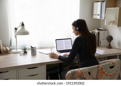 Back view of young Caucasian female student sit at desk at home study distant on laptop writing in notebook. Millennial woman in headphones work online or take course on internet. Education concept.