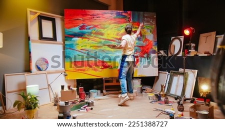 Back view of young Caucasian man painter emotionally drawing on big canvas using paint brush in cozy workshop creating abstract oil painting, contemporary fine art painter, creativity concept