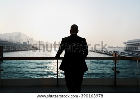 Back view of a young confident man financier is thinking about something, while is standing outdoors against sea port. Silhouette of male intelligent managing director is resting after hard work day