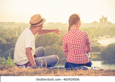 Back View Of Young Couple Looking At View And Enjoying The Sunset While Sitting On Hill Meadow Above The City. Summer And Love Concepts. 