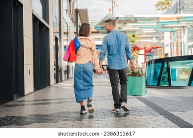 Back view of young couple shopping on a mall, carrying paper bags in their hands and walking together looking each other and smiling. Husband and wife sightseeing holding hands enjoying on a date - Powered by Shutterstock